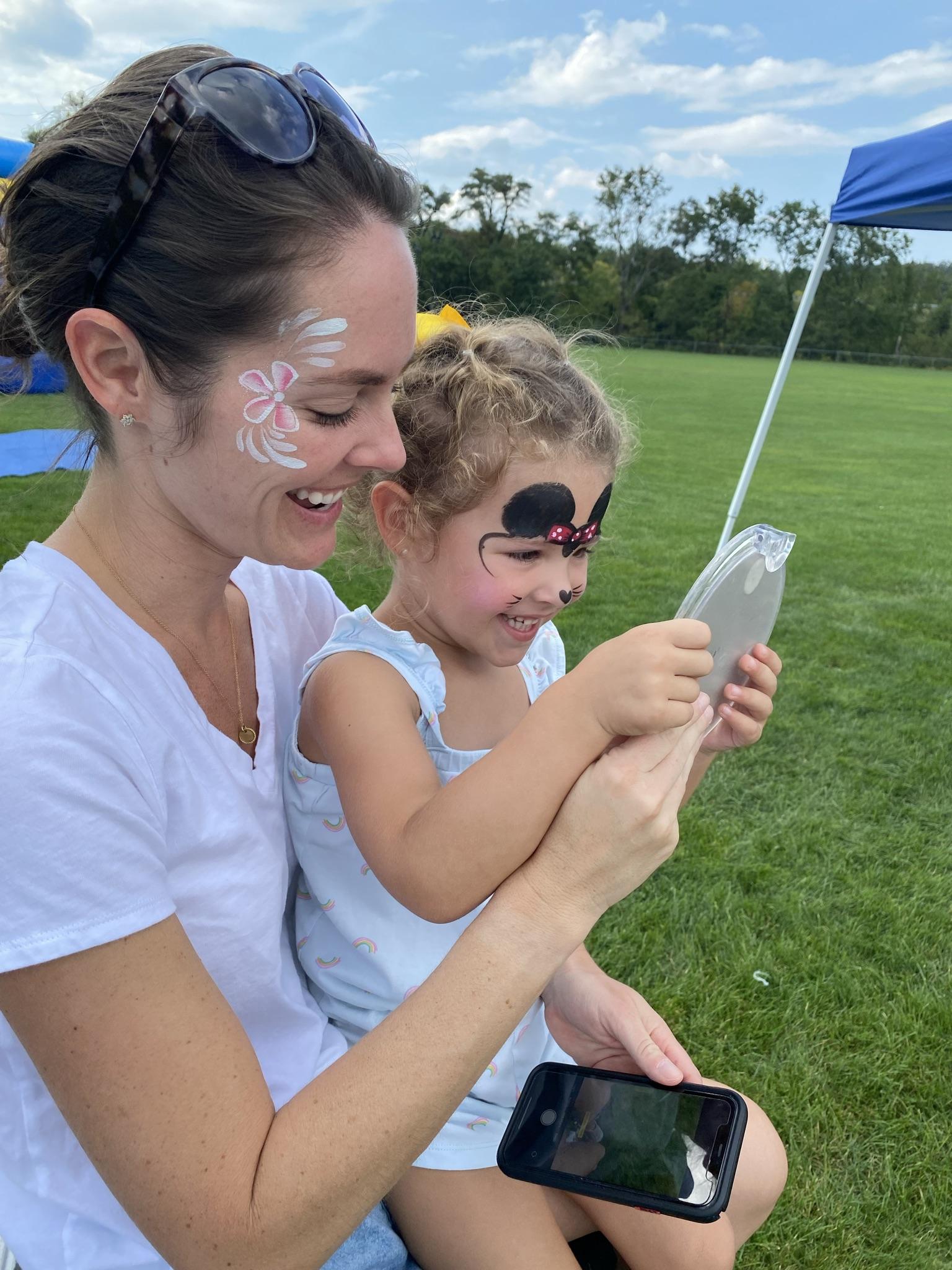 Mom and Daughter painted to like like Minnie Mouse looking into a small mirror to see the painting